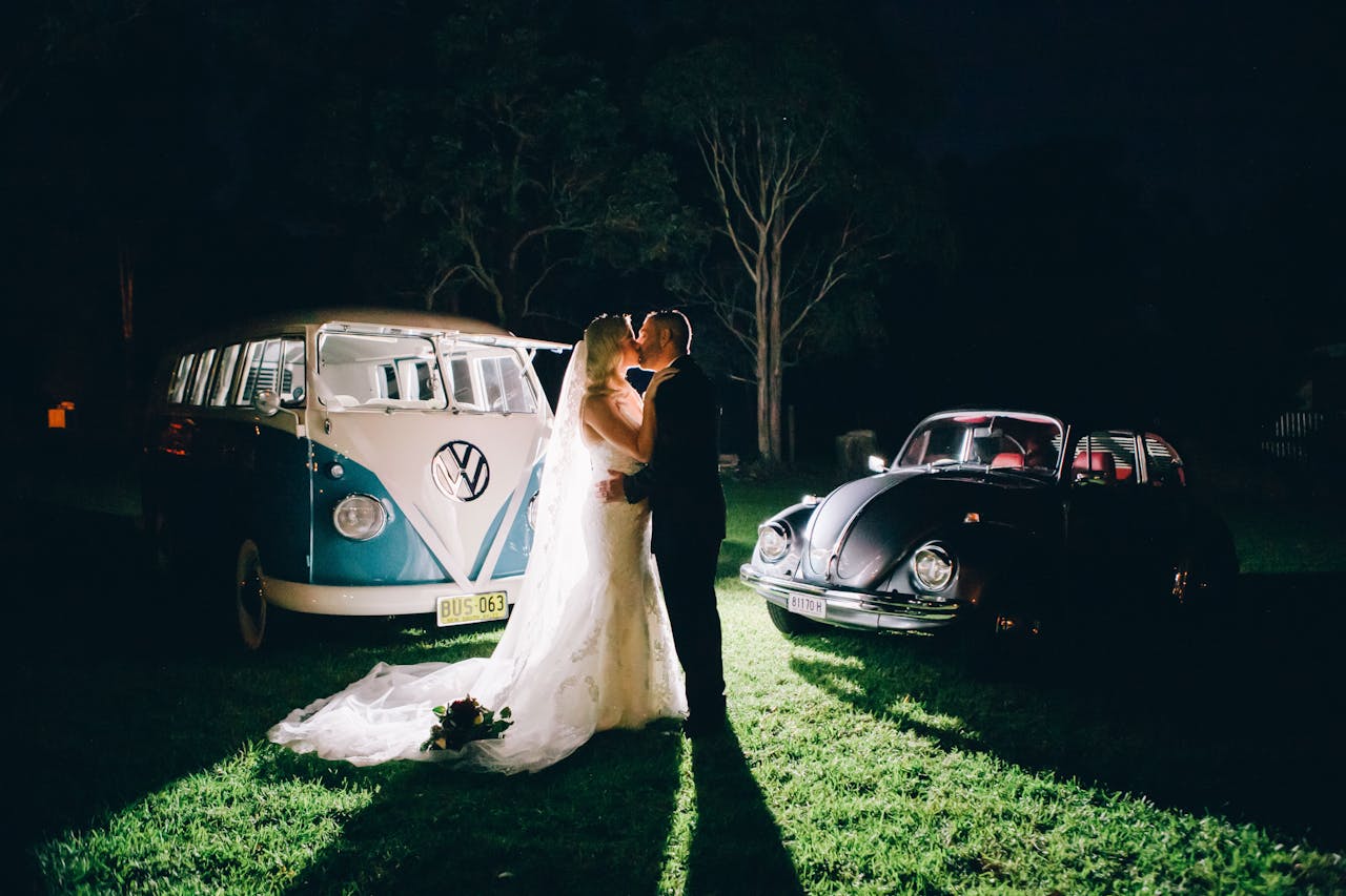 A bride and groom share a kiss beside vintage VW cars during a rustic nighttime wedding in Australia.